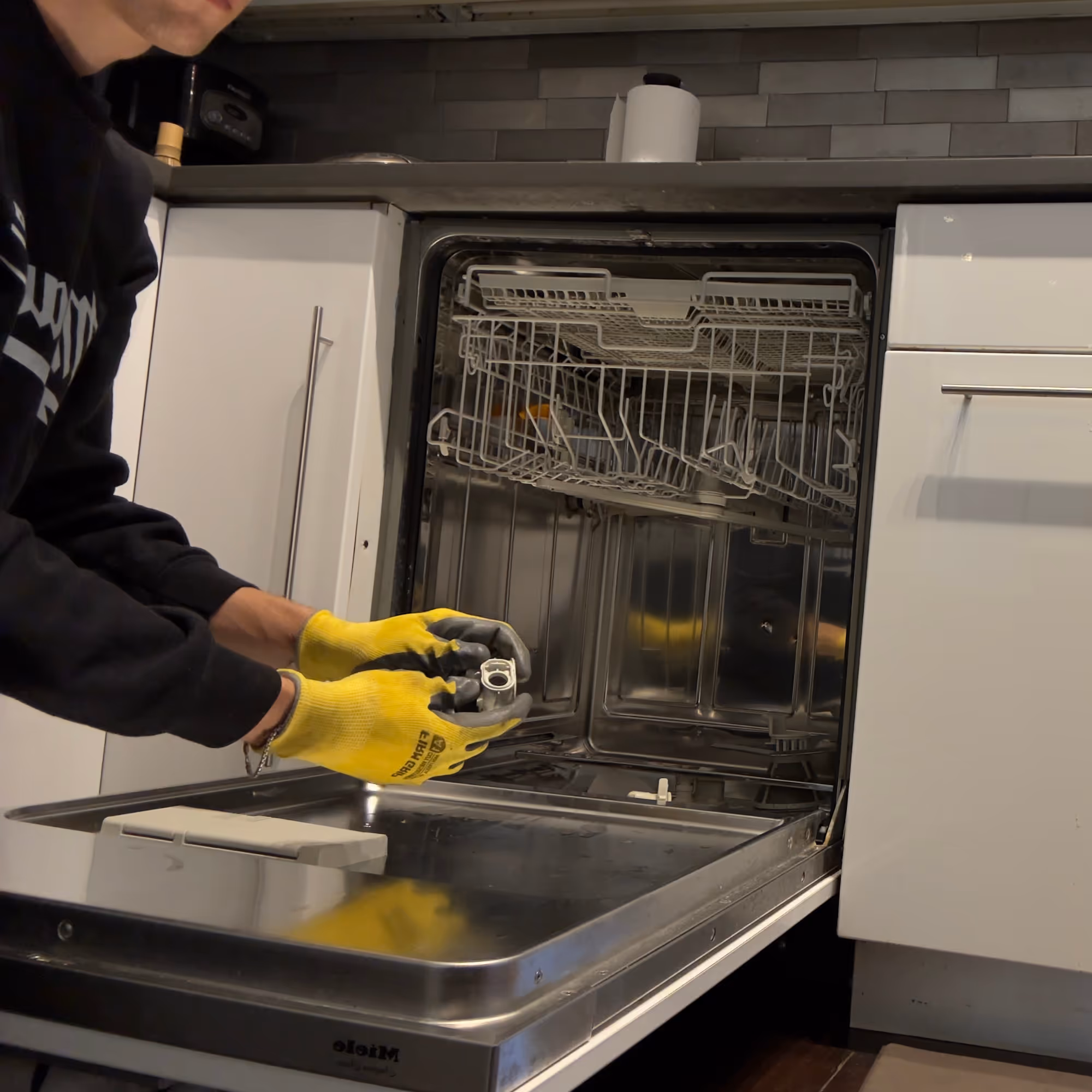 Person wearing yellow gloves holding a dishwasher filter above an open Miele dishwasher in a kitchen.