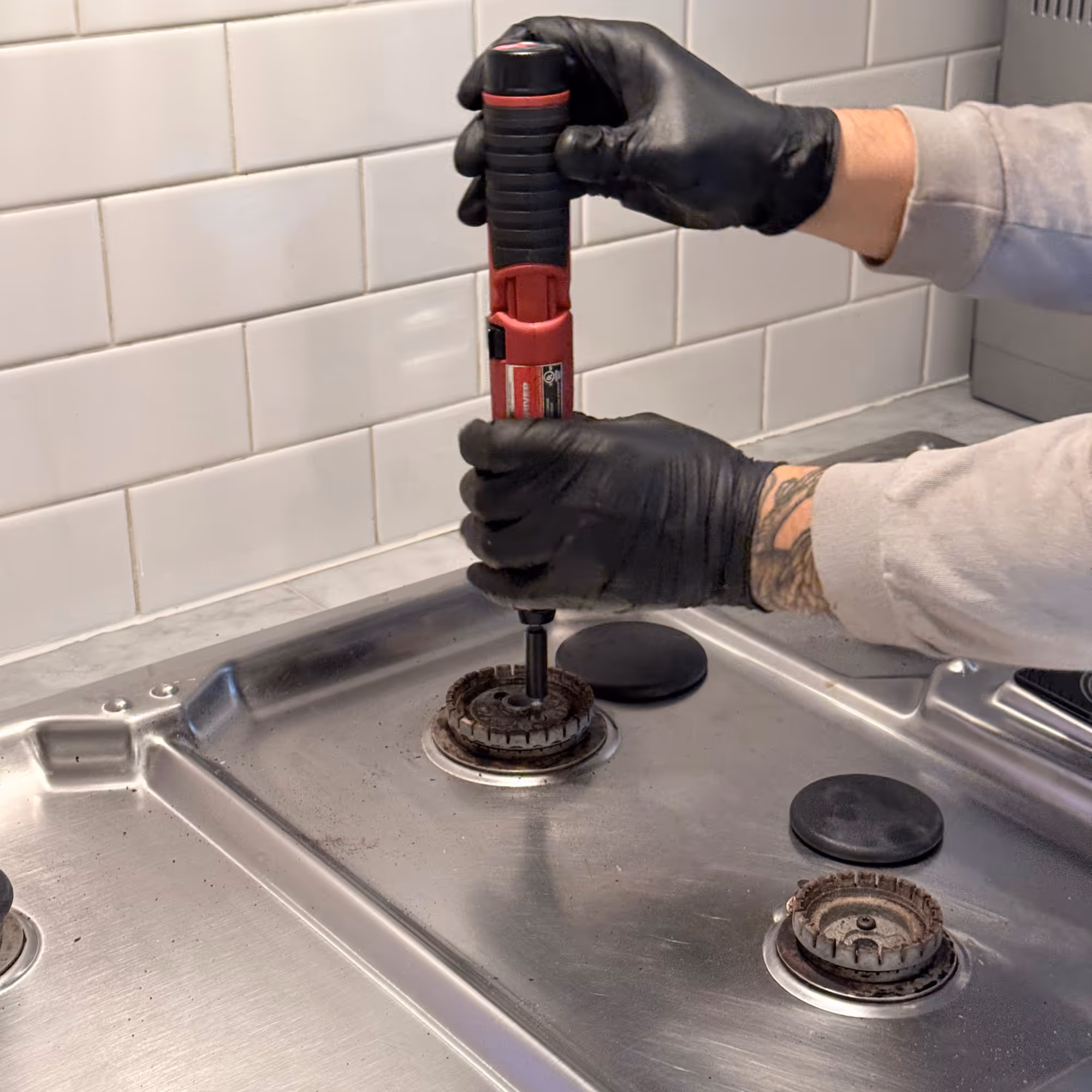 Person wearing black gloves using a red screwdriver to repair a gas stove burner on a stainless steel cooktop.