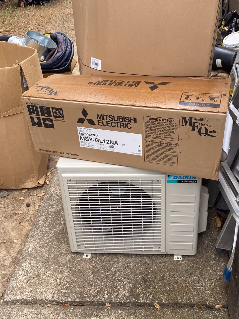 Daikin outdoor air conditioning unit with a Mitsubishi Electric cardboard box on top, placed on a concrete surface near other cardboard boxes and equipment.