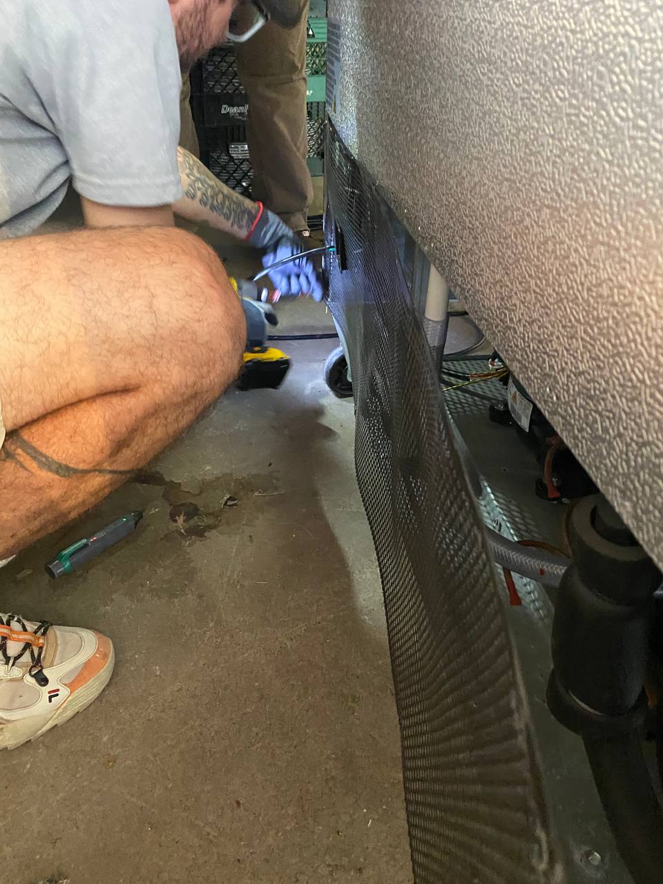 Person wearing gloves crouches while using a tool to repair or inspect the side of a large appliance or machine.