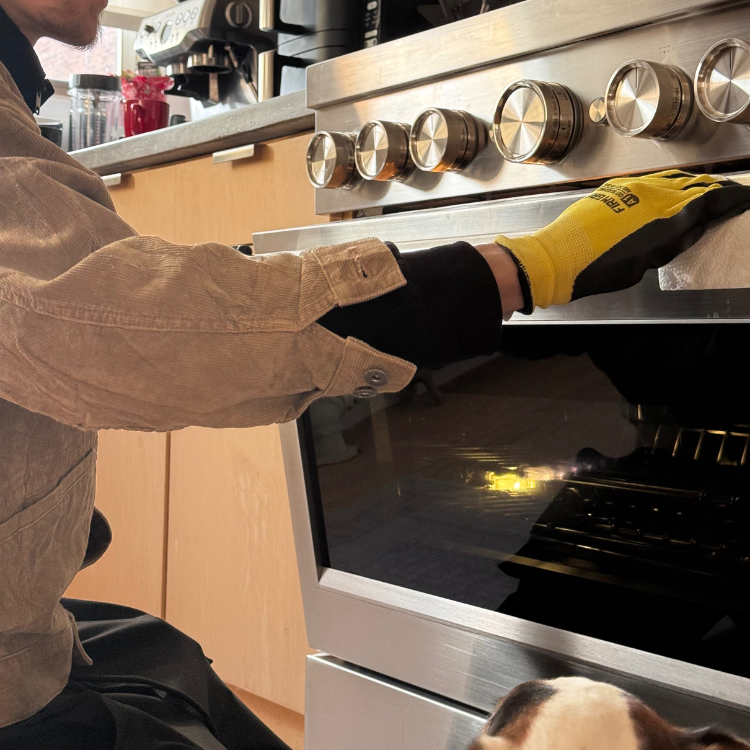 Person wearing a yellow and black glove adjusting a stainless steel oven knob with a dog looking up in the kitchen.