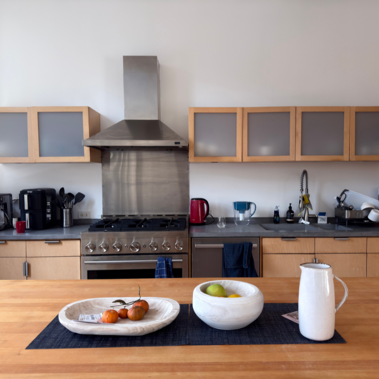 Modern kitchen with wooden cabinets, stainless steel stove and hood, countertop appliances, and a wooden table with two bowls of fruit and a white pitcher.