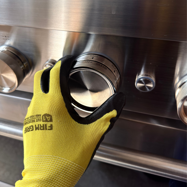 Hand wearing a yellow and black work glove gripping a stainless steel oven control knob.