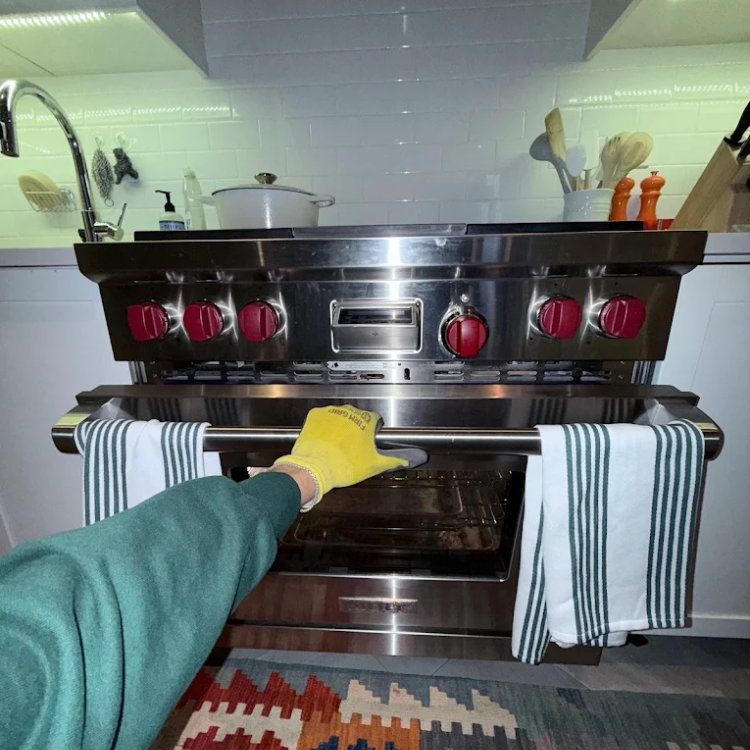 Person wearing a green sleeve and yellow oven mitt opening a stainless steel oven door with red knobs in a modern kitchen.