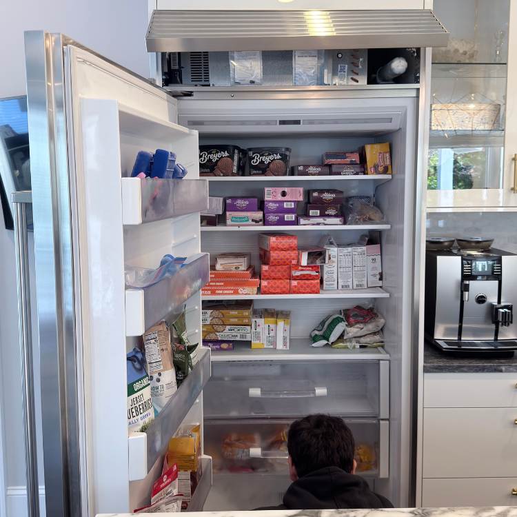 Open freezer stocked with various food items including ice cream, boxes, and bags, with a person kneeling in front of it in a modern kitchen.