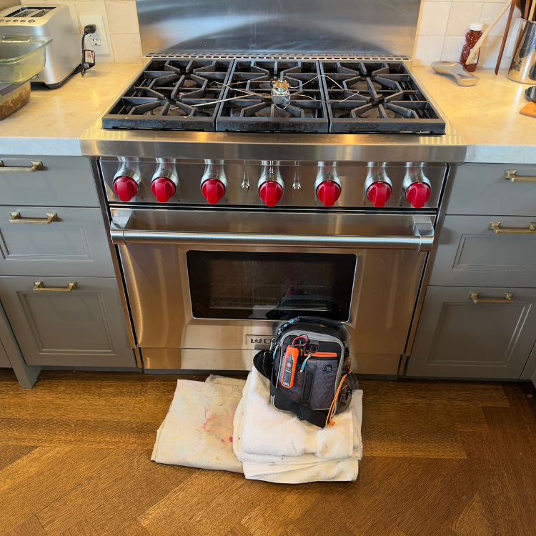 Modern kitchen stove with red knobs and a tool bag on folded towels placed on the wooden floor in front of it.