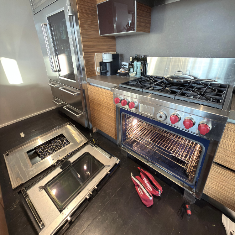 Modern kitchen with a stainless steel gas stove with five burners and an open oven door detached and lying on the floor, alongside a tool bag and screwdriver.
