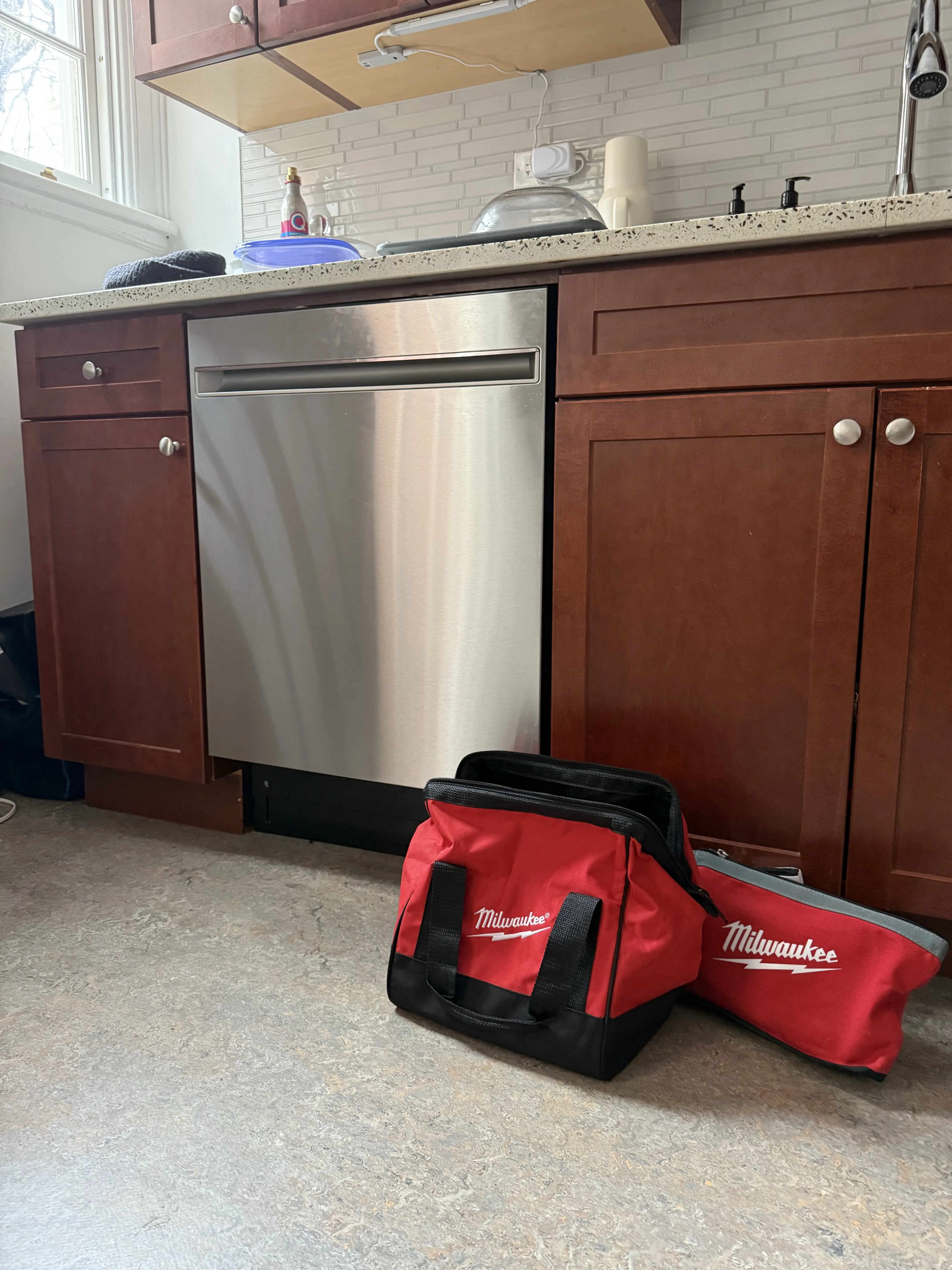 Red Milwaukee tool bags on kitchen floor near stainless steel dishwasher