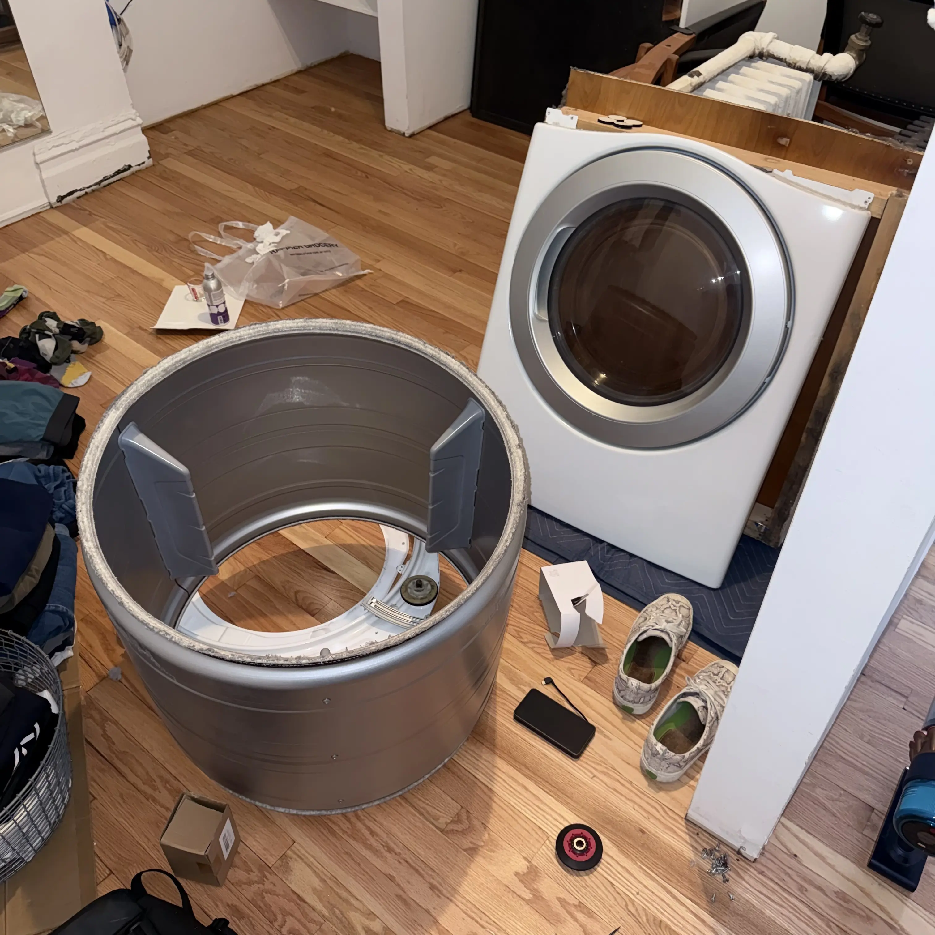 Disassembled washing machine drum on floor with washer and clothes nearby