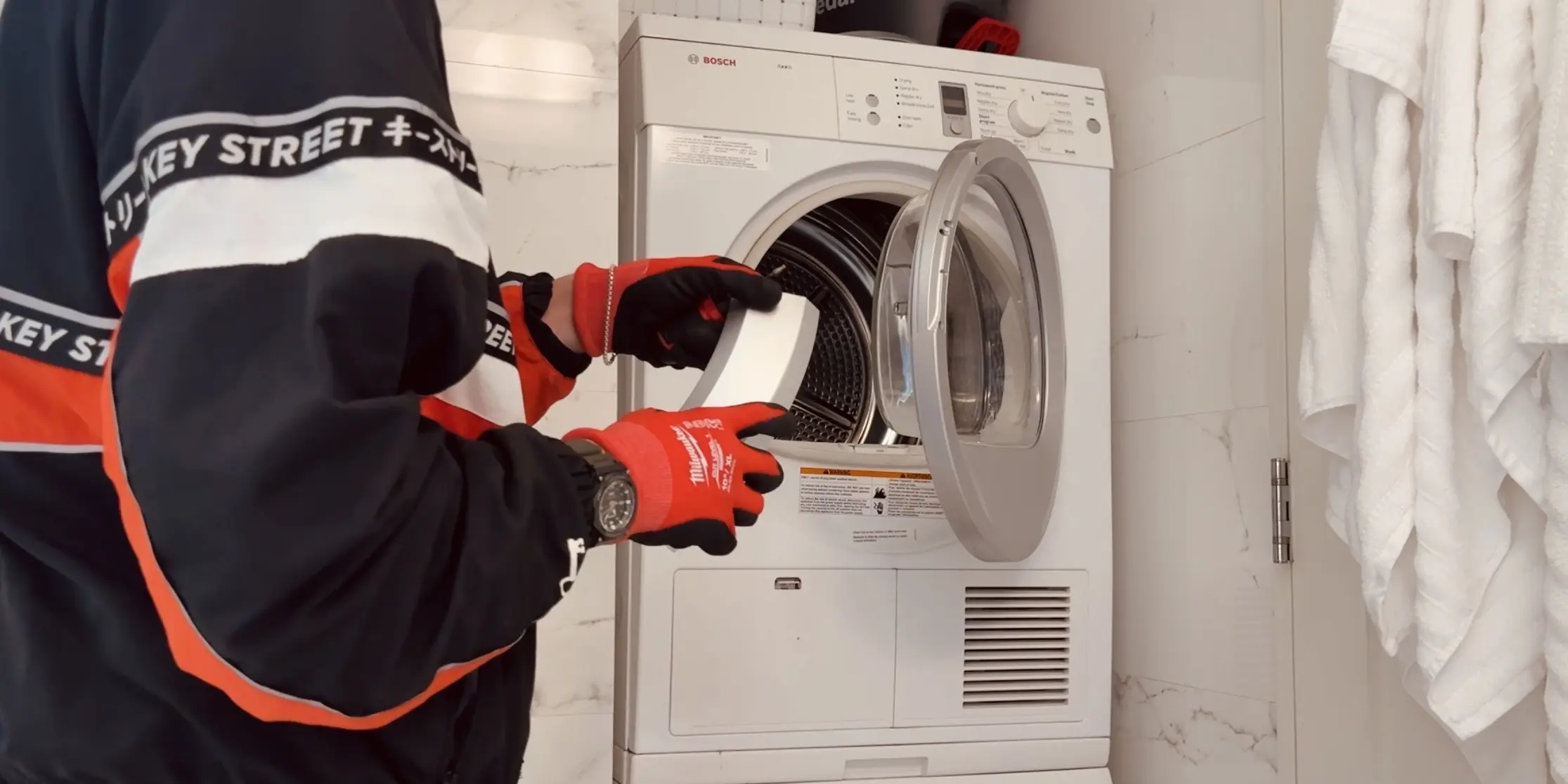 Modern kitchen corner featuring a stainless steel glass-door refrigerator, wooden cabinets, a mounted flat-screen TV, and a coffee machine on the countertop.