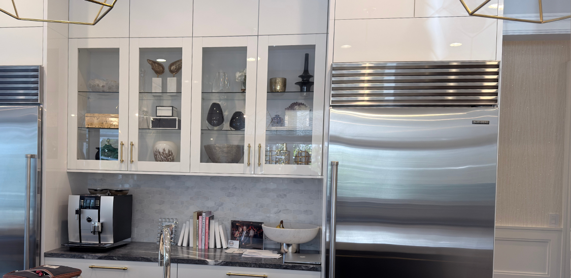Modern kitchen corner featuring a stainless steel glass-door refrigerator, wooden cabinets, a mounted flat-screen TV, and a coffee machine on the countertop.