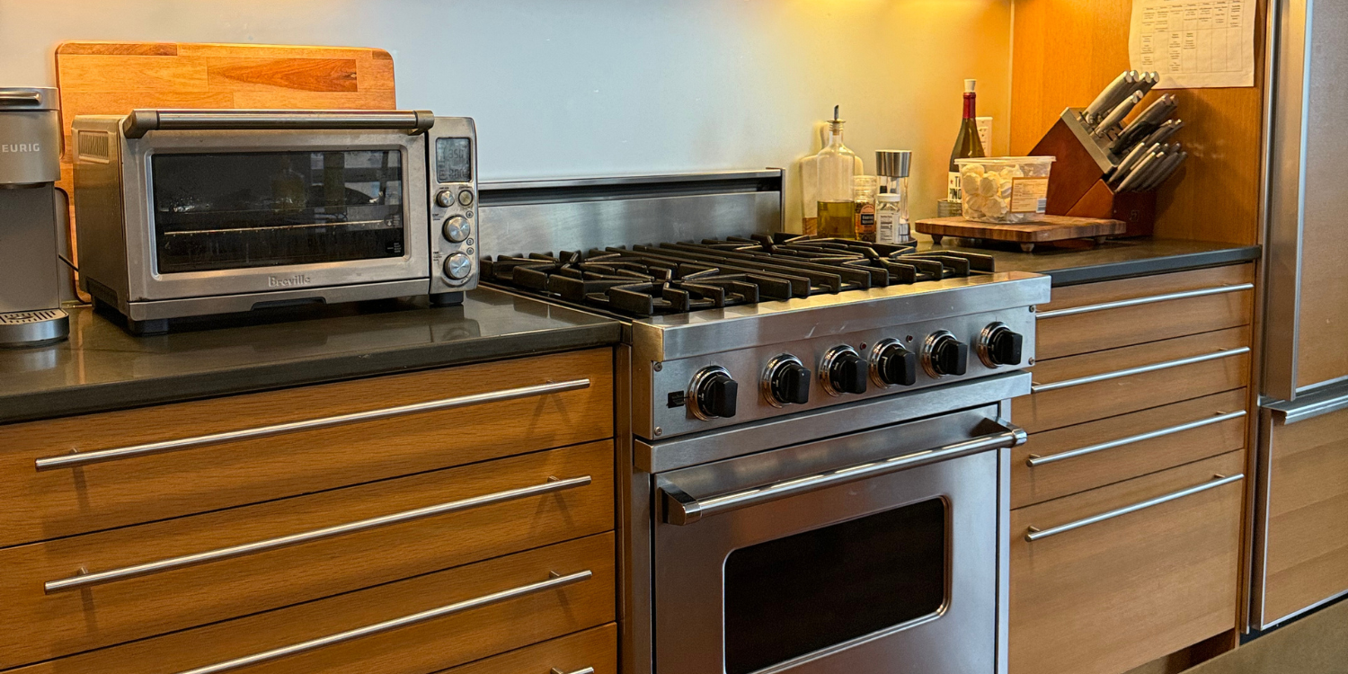 Modern kitchen corner featuring a stainless steel glass-door refrigerator, wooden cabinets, a mounted flat-screen TV, and a coffee machine on the countertop.