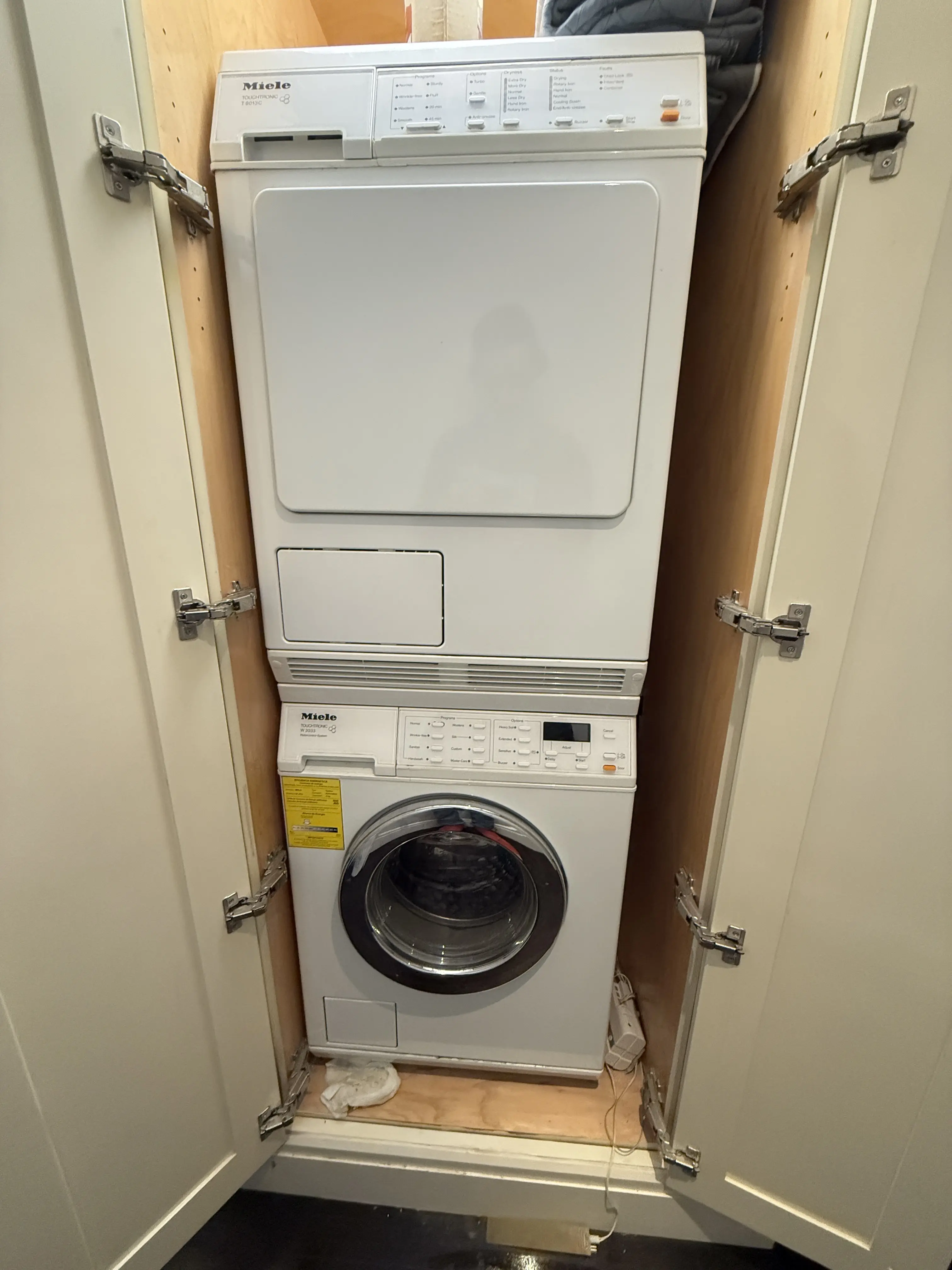 Modern kitchen corner featuring a stainless steel glass-door refrigerator, wooden cabinets, a mounted flat-screen TV, and a coffee machine on the countertop.