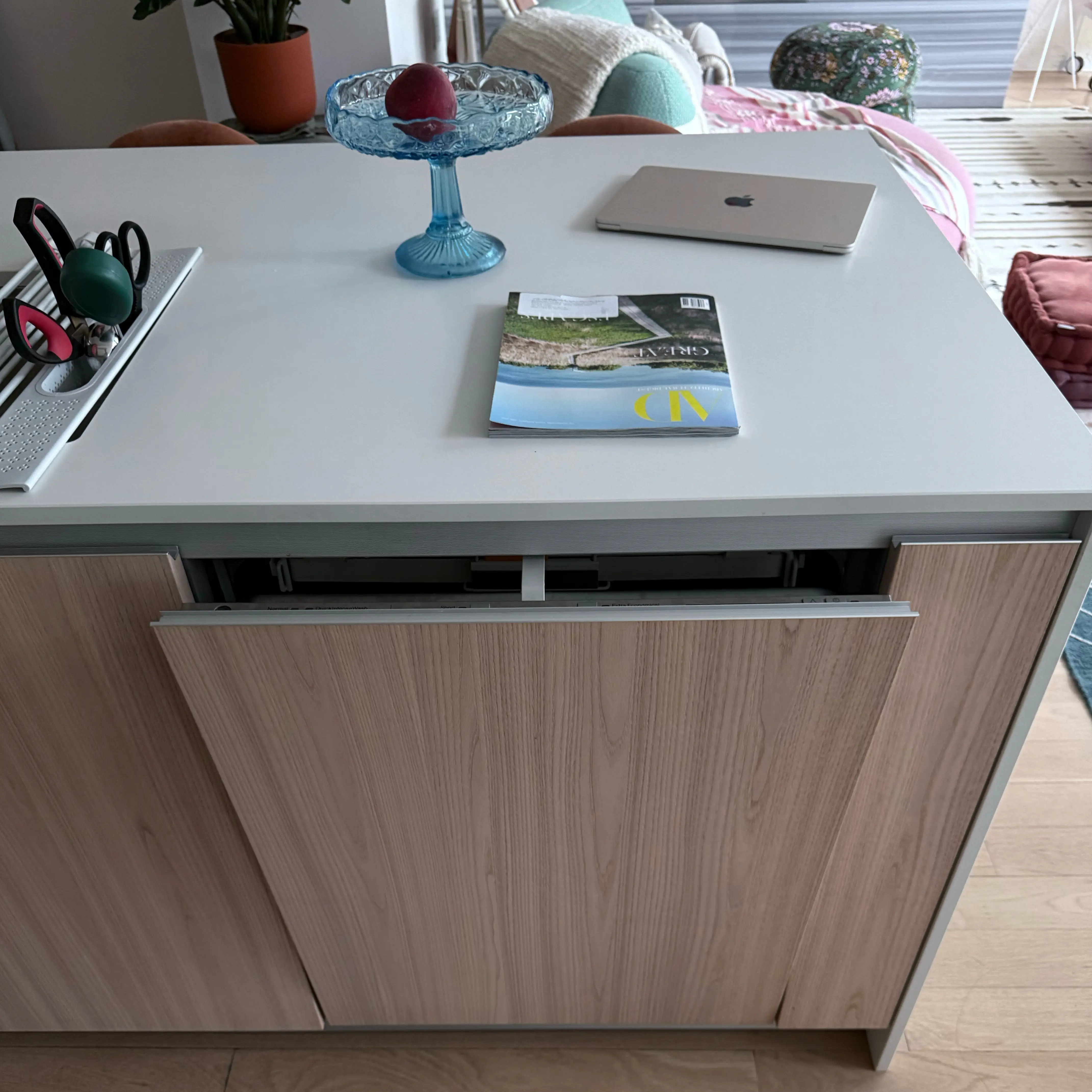 Modern kitchen corner featuring a stainless steel glass-door refrigerator, wooden cabinets, a mounted flat-screen TV, and a coffee machine on the countertop.