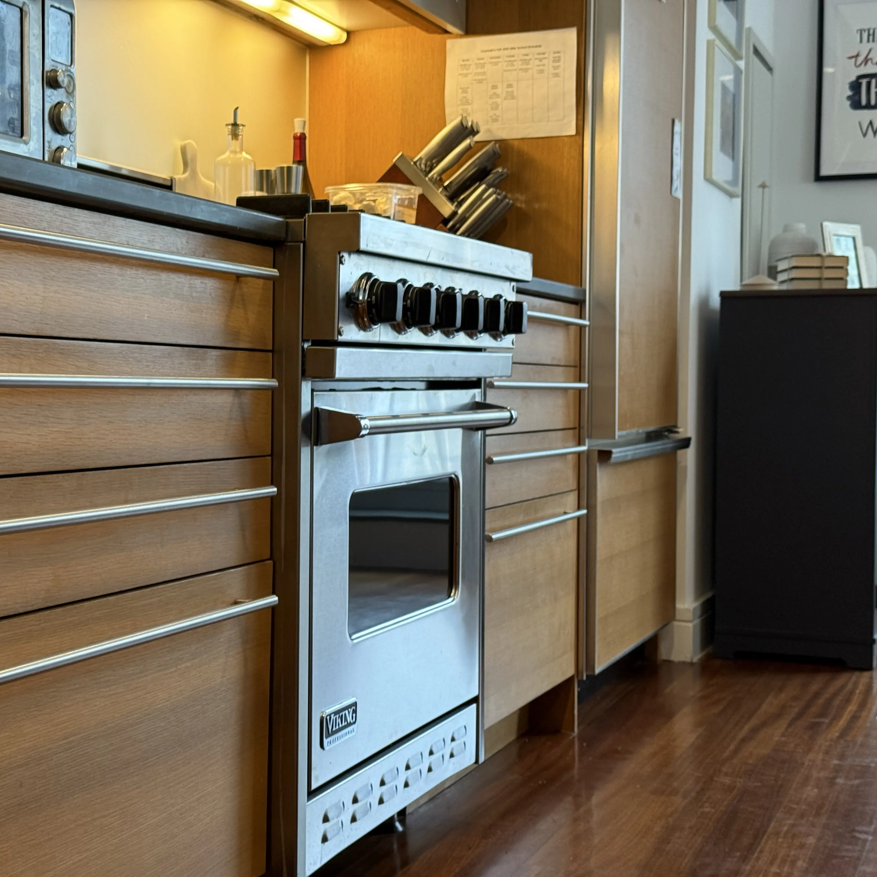 Modern kitchen corner featuring a stainless steel glass-door refrigerator, wooden cabinets, a mounted flat-screen TV, and a coffee machine on the countertop.