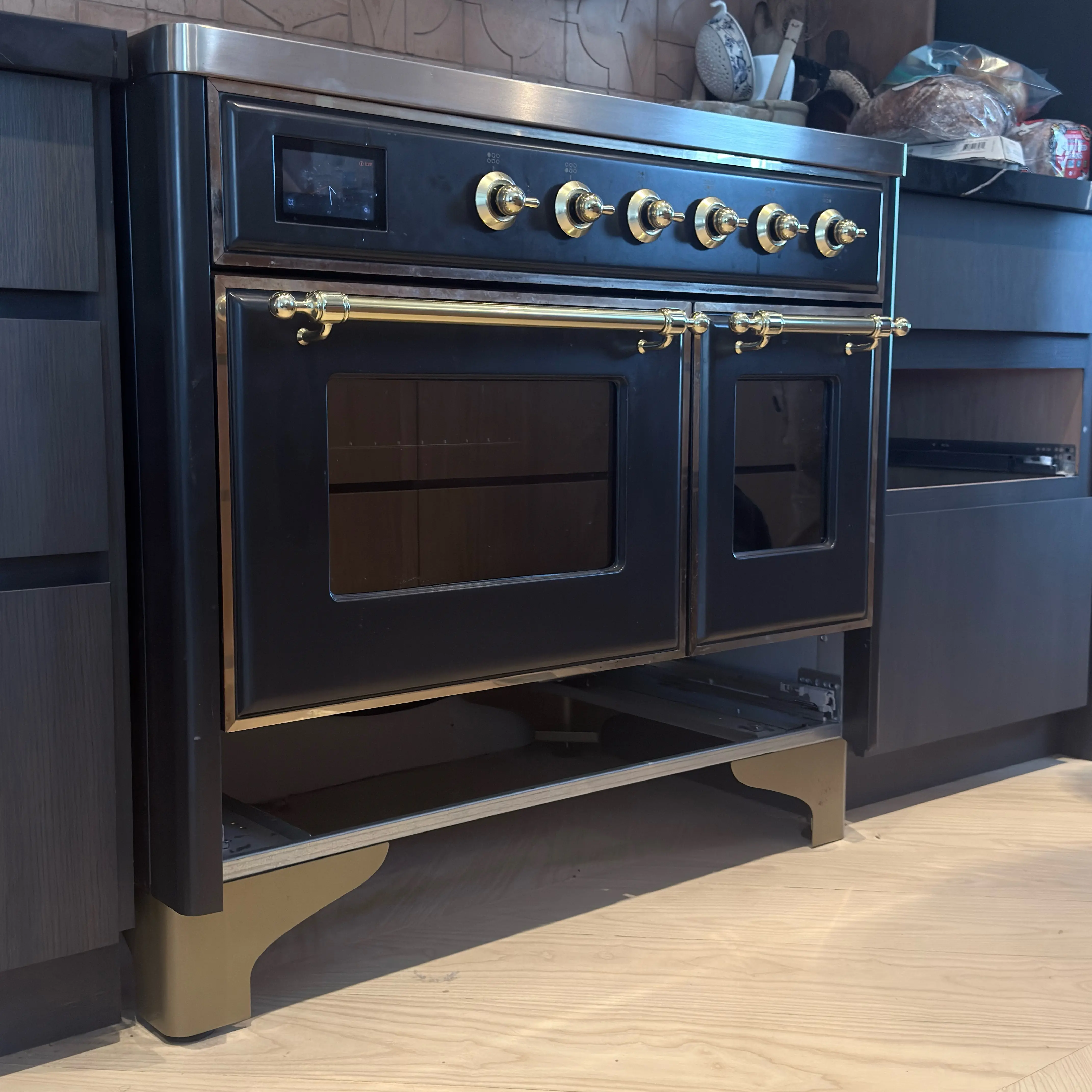 Modern kitchen corner featuring a stainless steel glass-door refrigerator, wooden cabinets, a mounted flat-screen TV, and a coffee machine on the countertop.
