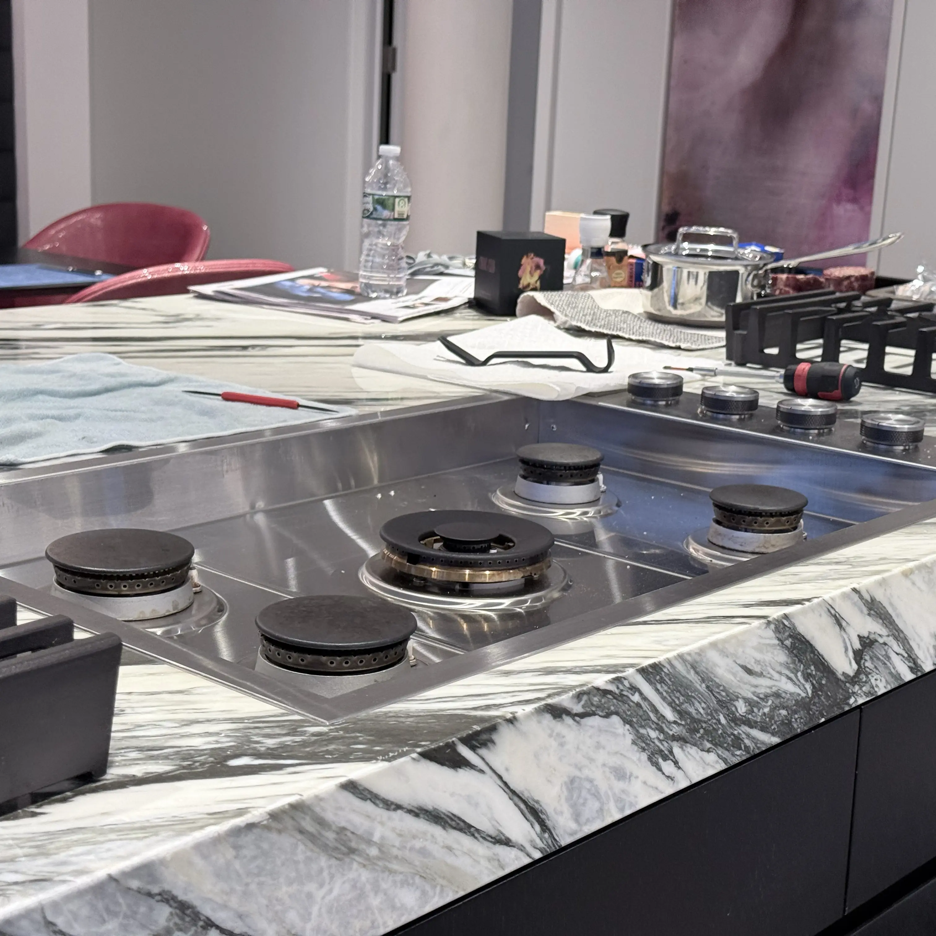Modern kitchen corner featuring a stainless steel glass-door refrigerator, wooden cabinets, a mounted flat-screen TV, and a coffee machine on the countertop.