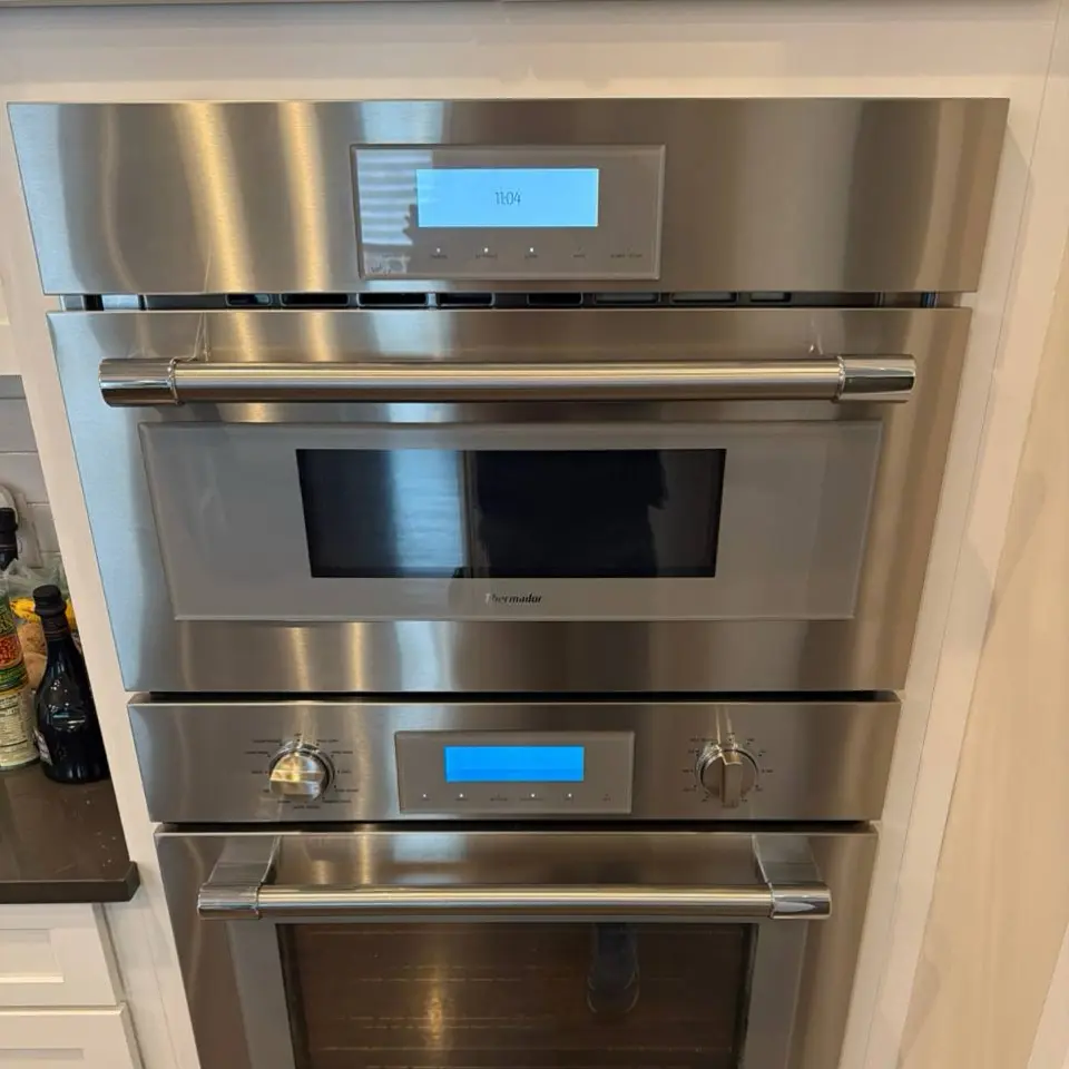 Modern kitchen corner featuring a stainless steel glass-door refrigerator, wooden cabinets, a mounted flat-screen TV, and a coffee machine on the countertop.