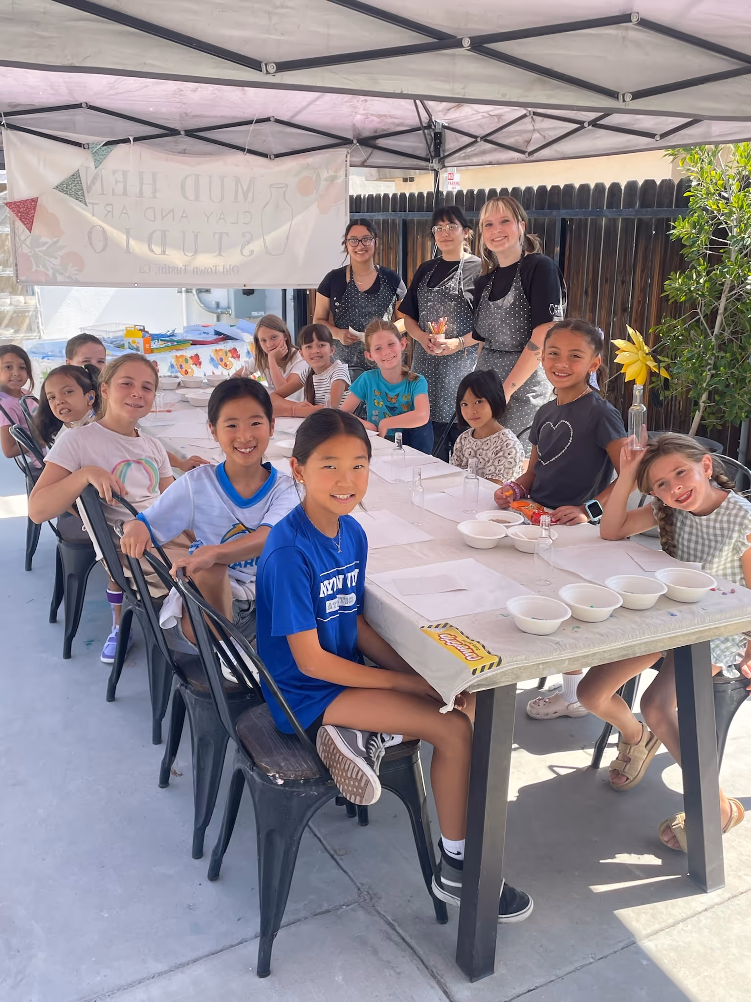 Group of children seated around a table under a canopy with adults standing behind them