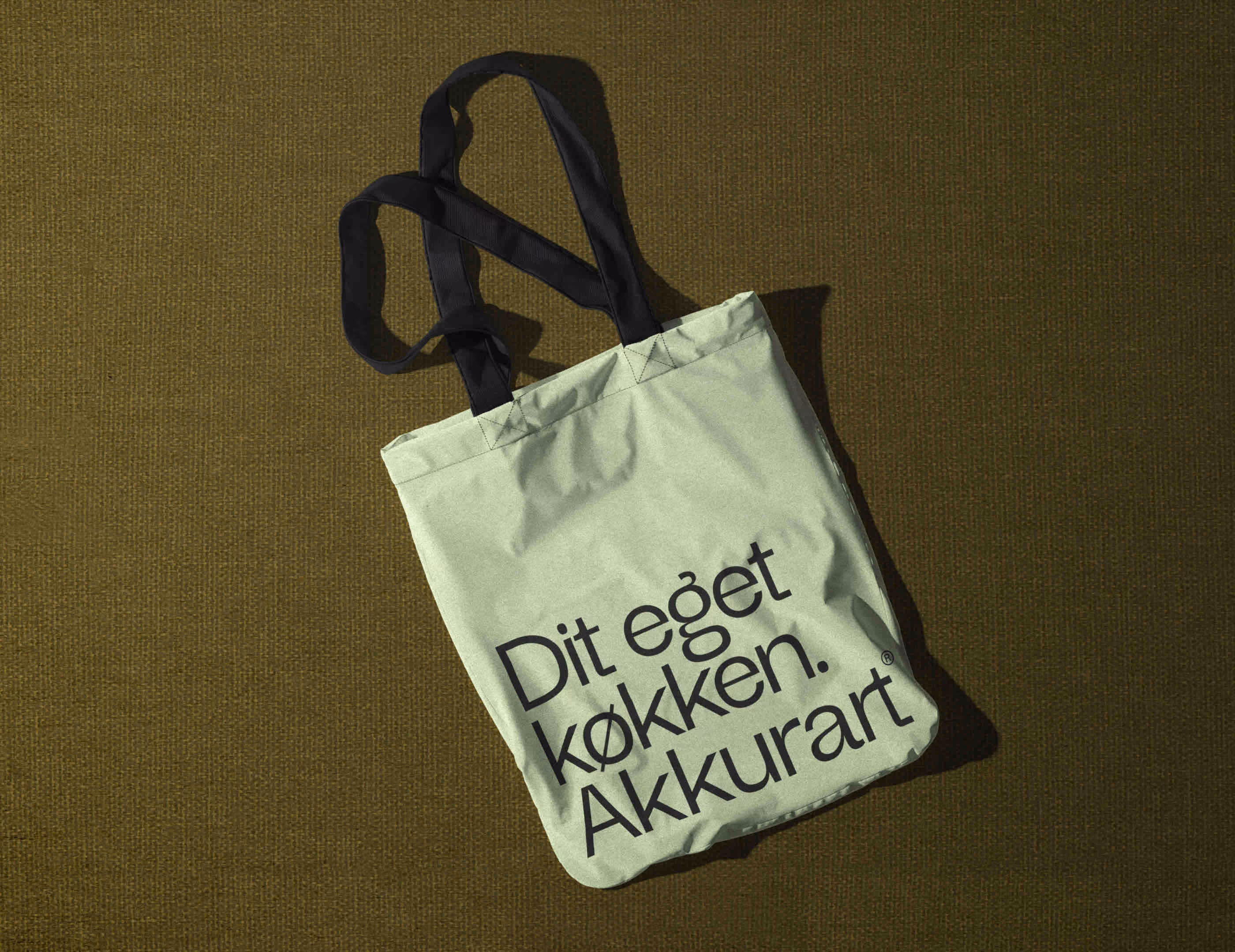 En lys mulepose med sort tekst 'Dit eget køkken. Akkurart' og sorte håndtag på en mørk stofbaggrund.