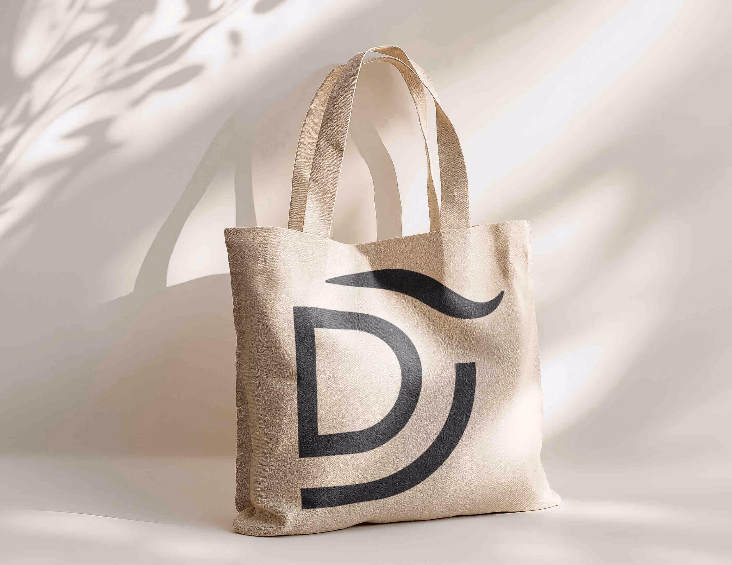 Beige stofindkøbstaske med sort bogstav D-logo, stående mod lys baggrund med skygger.