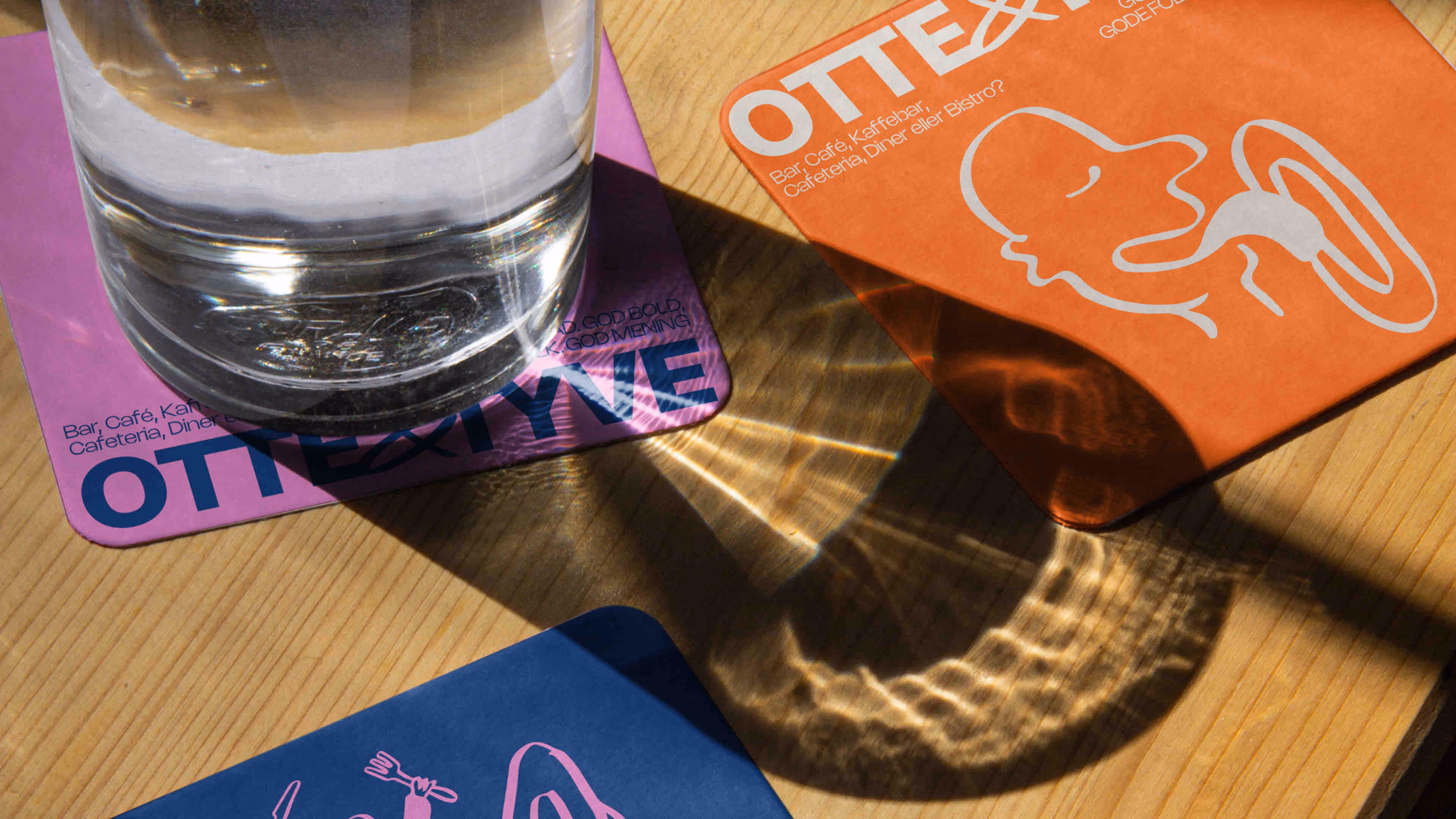 Et glas med vand står på en lilla coaster med teksten OTTETYE, ved siden af en orange coaster med et stiliseret hvidt ansigt, og en blå coaster delvist synlig nedenfor på et lyst træbord.