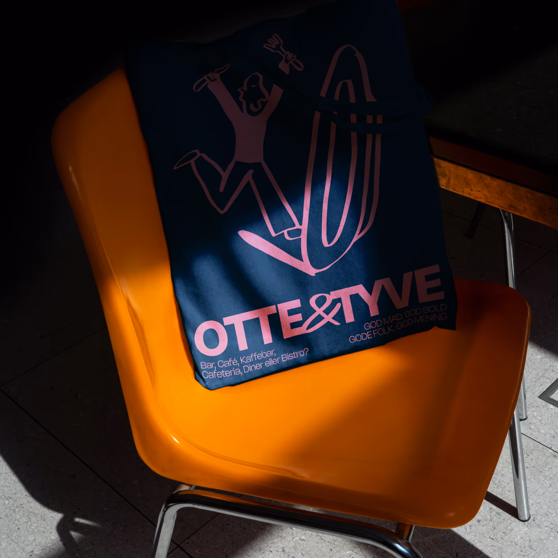 Mørkeblå taske med pink tekst og illustration hængende på en orange stol med metalben.