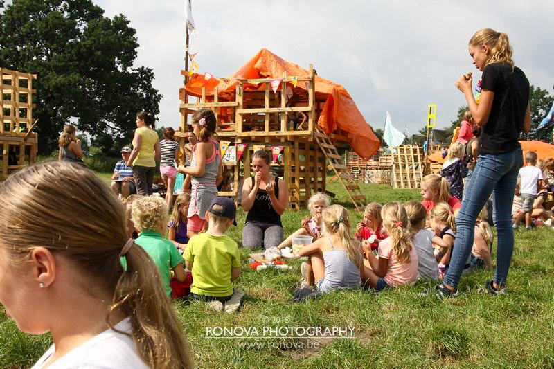 Foto Doe Week terrein met zelfgebouwde hutten en groepen kinderen die genieten in het gras of aan het spelen zijn.