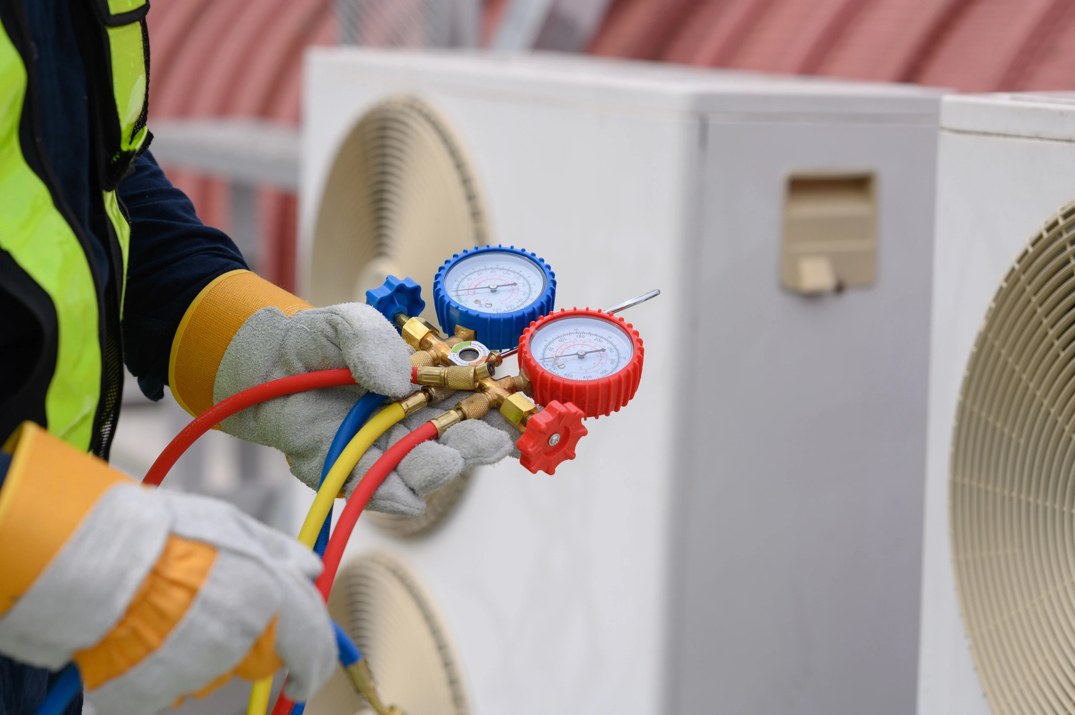 A technician wearing gloves and a safety vest adjusts HVAC gauges with colorful hoses, in front of large air conditioning units.
