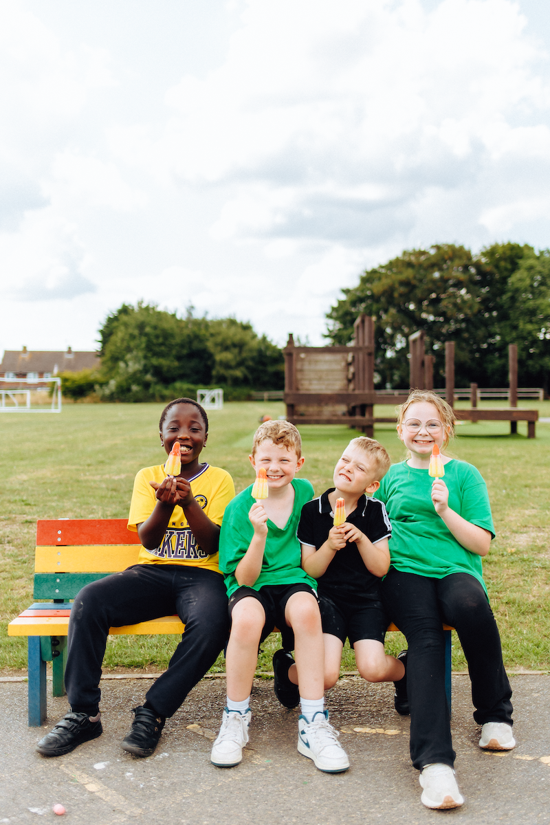 Four children sitting on a colorful bench in a park, smiling and ice lollies on a sunny day.