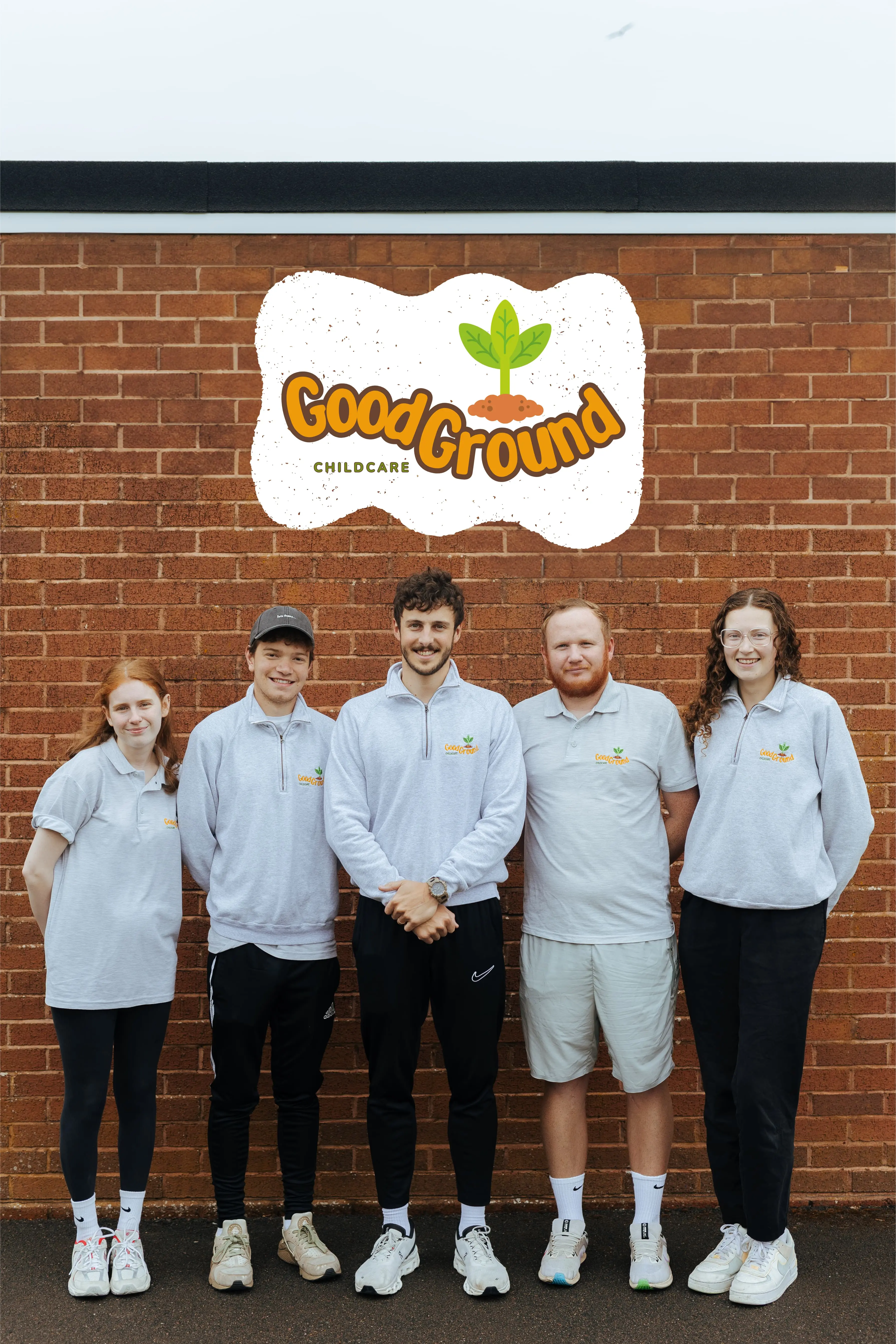 Five young adults wearing light gray GoodGround Childcare shirts standing in front of a brick wall with the GoodGround Childcare logo above them.