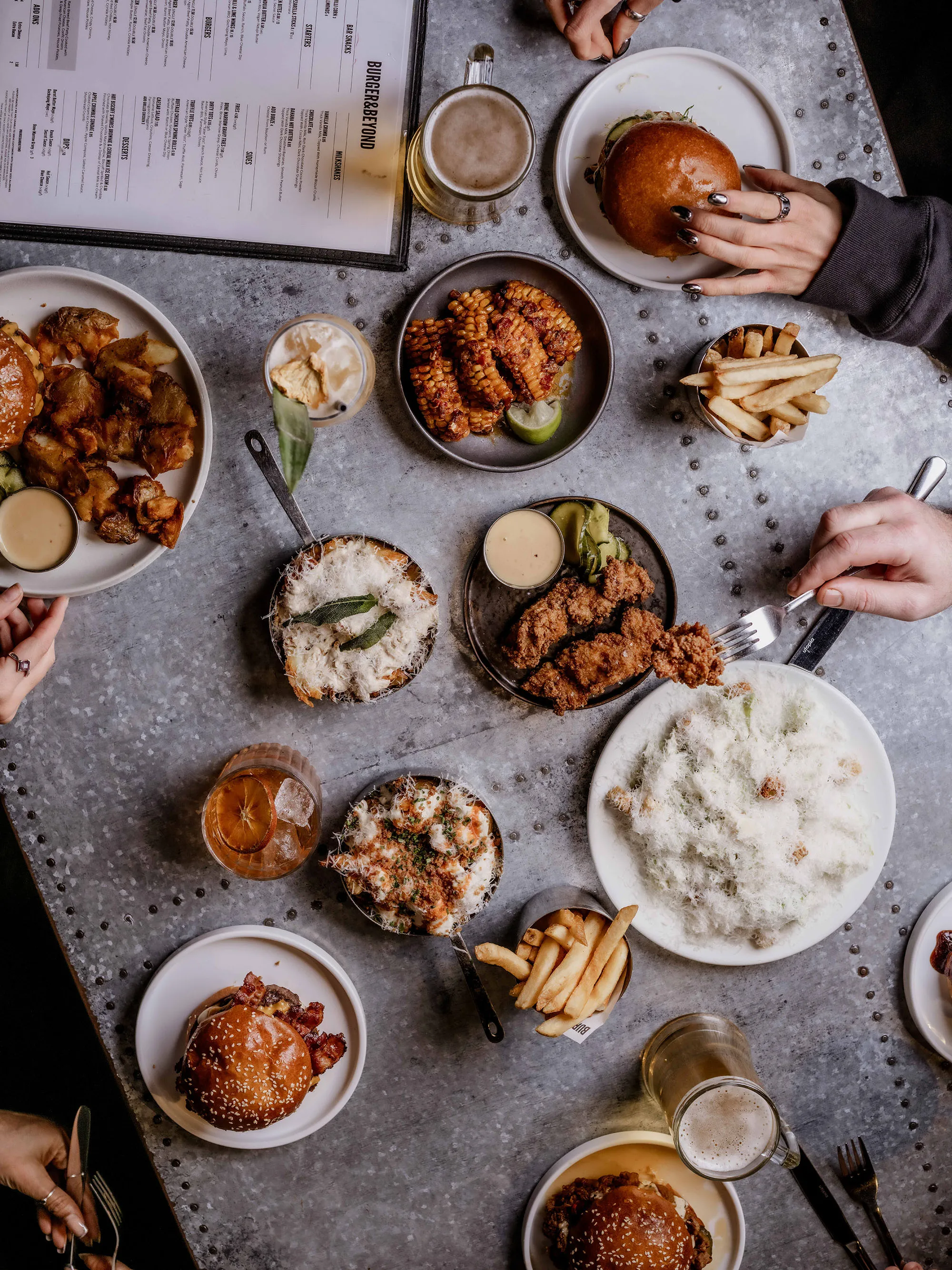Overhead view of a table with burgers, fried chicken, grilled corn, fries, drinks, and a menu with people reaching for food.