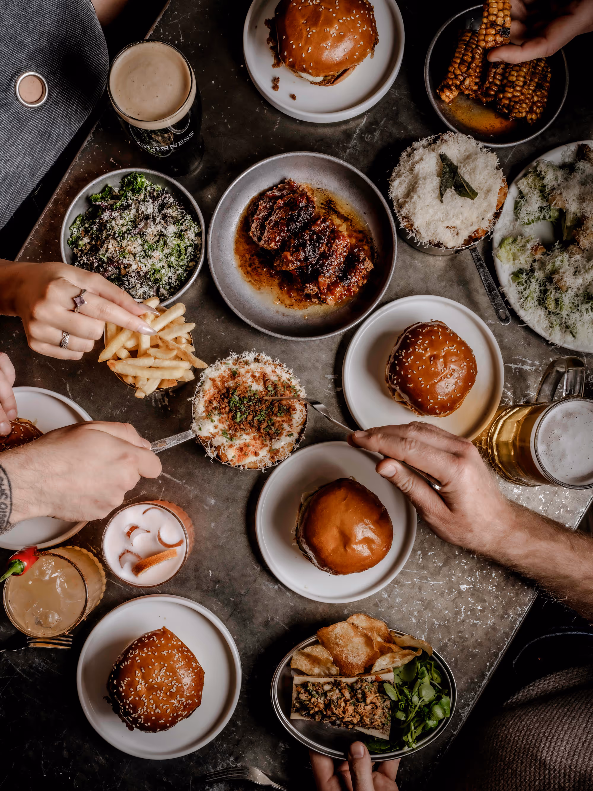 Top-down view of a table with burgers, fried chicken tenders, fries, grilled corn, cheesy mashed potatoes, and drinks with hands reaching for the food.