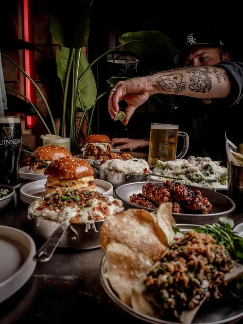 Top-down view of a table with burgers, fried chicken tenders, fries, grilled corn, cheesy mashed potatoes, and drinks with hands reaching for the food.