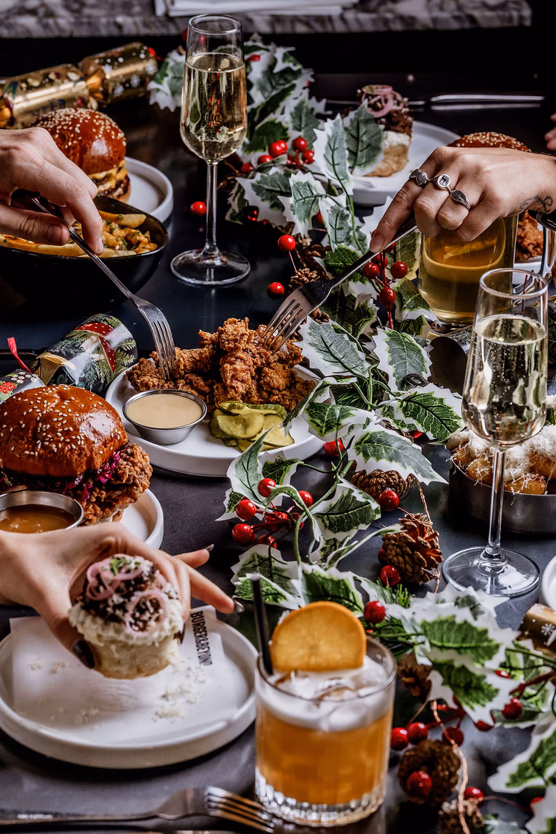 Hands reaching for fried chicken, cupcake, burgers, and drinks on a festive table with holly and pinecones.