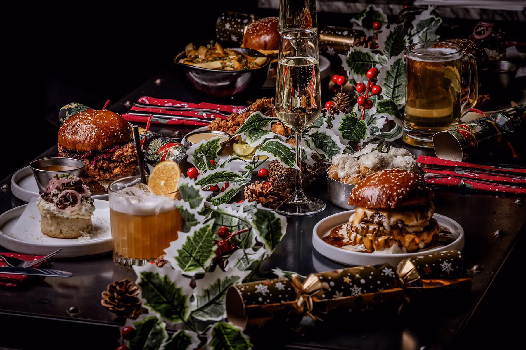 Festive table with burgers, chicken wings, assorted drinks, and holiday decorations including holly and pine cones.