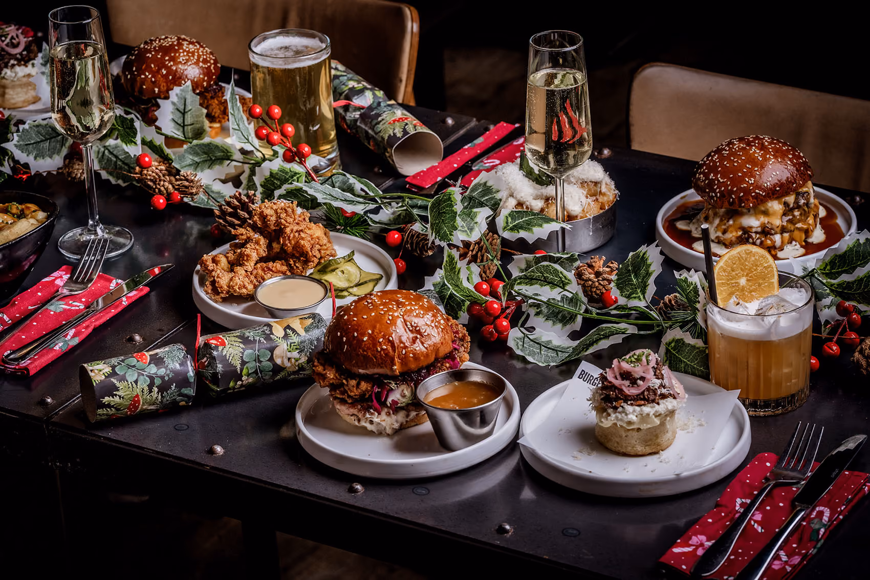 Festive table with burgers, fried chicken, desserts, drinks, and holiday decorations with holly and pine cones.