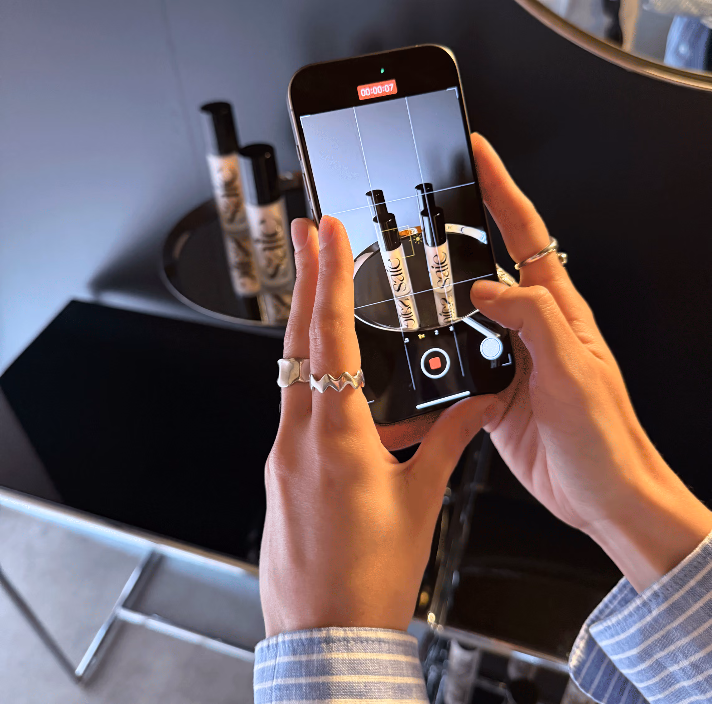 Hands holding smartphone recording two cosmetic bottles on a black round tray with rings on fingers.