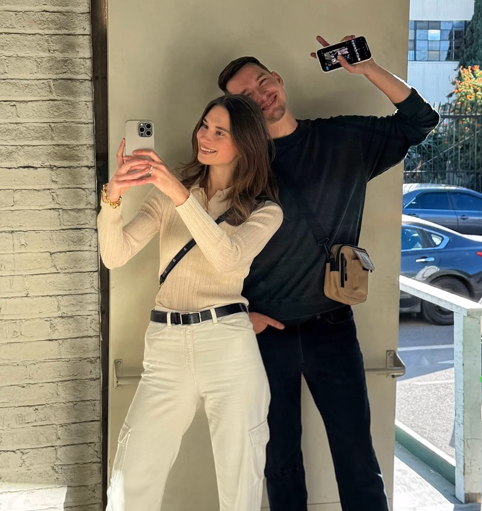 A smiling woman takes a selfie with a man behind her, who holds up his phone and flashes a peace sign.