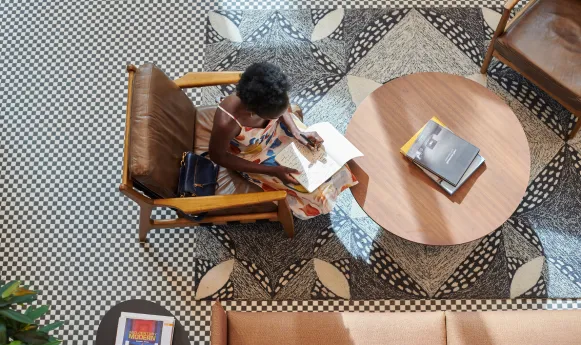 A woman sits in a stylish lounge, reading and writing on a patterned floor with modern decor.