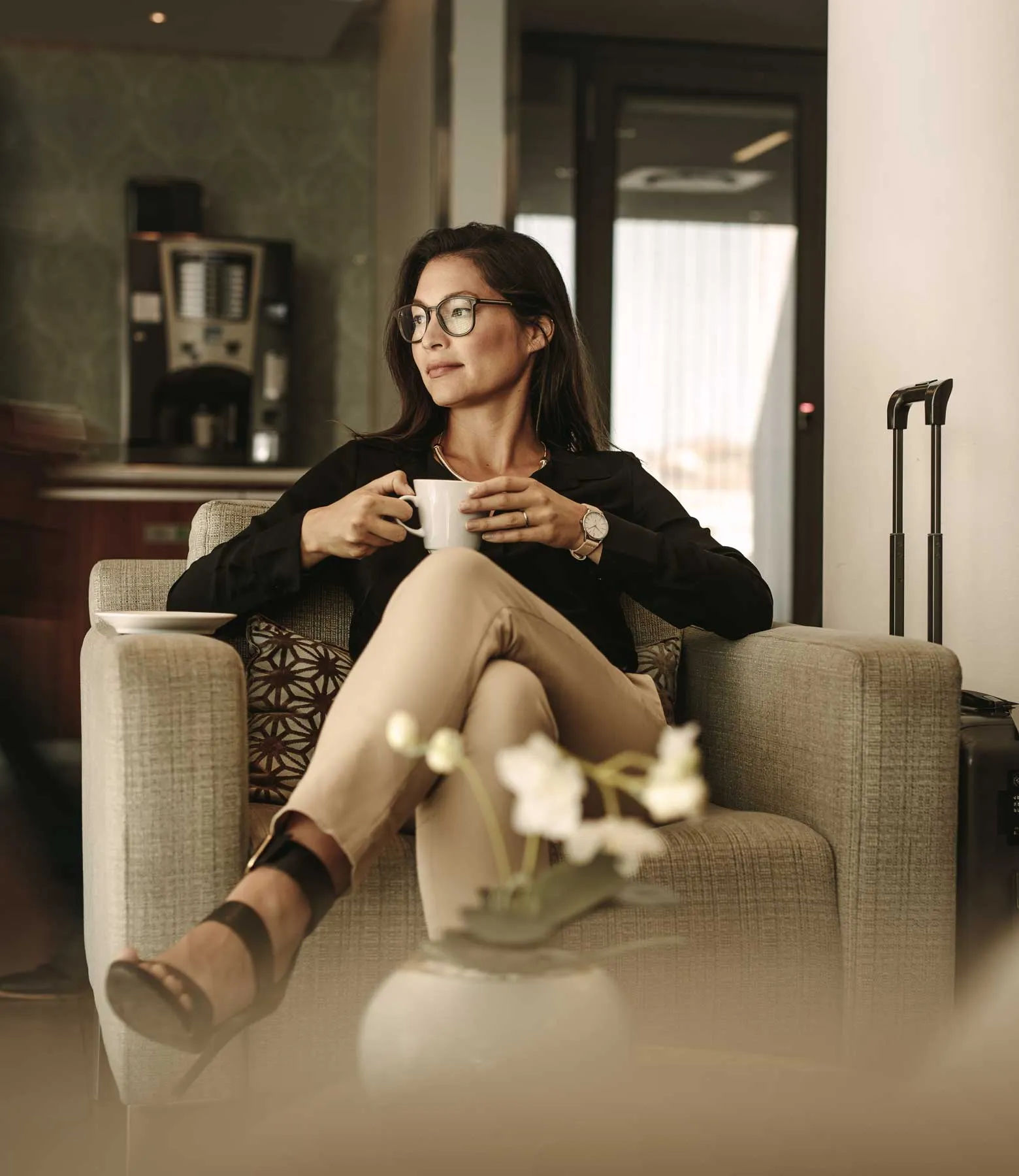 A woman sits in a lounge chair holding a cup, dressed in business attire with luggage nearby.