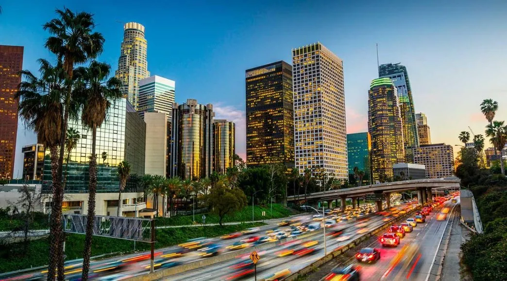 Downtown Los Angeles skyline at dusk with illuminated buildings and busy freeway traffic.