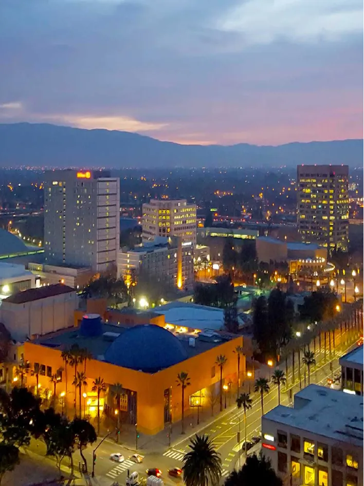 A vibrant cityscape at dusk featuring illuminated buildings, palm-lined streets, and distant hills.
