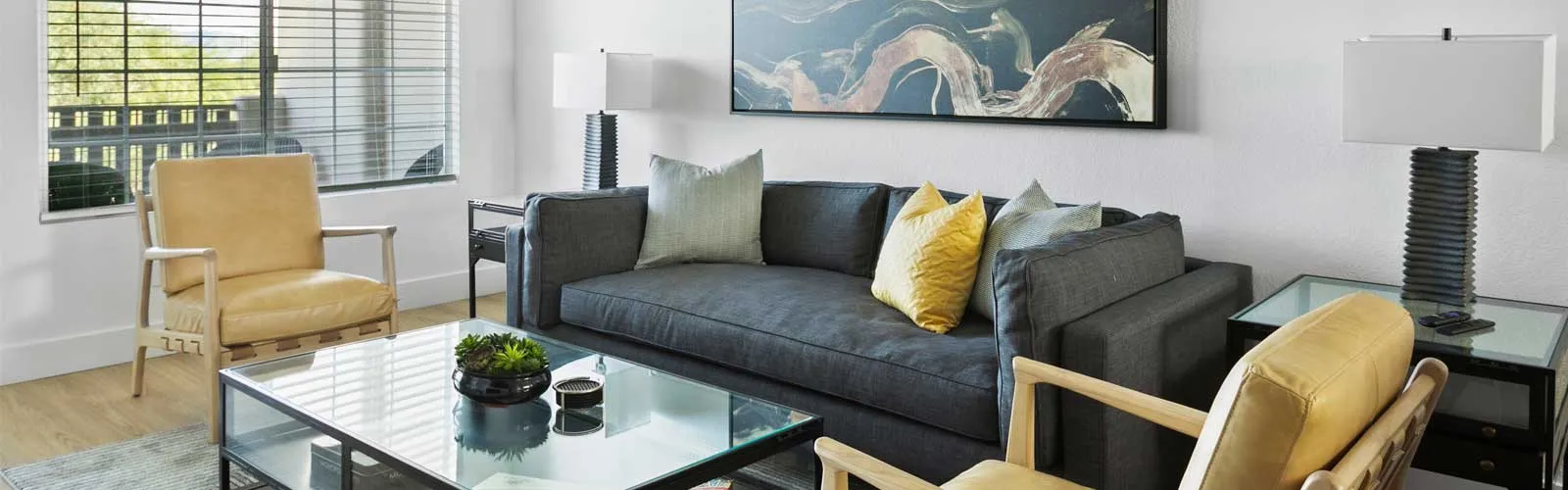 Modern living room with a dark sofa, yellow chairs, abstract art, and glass-topped tables.
