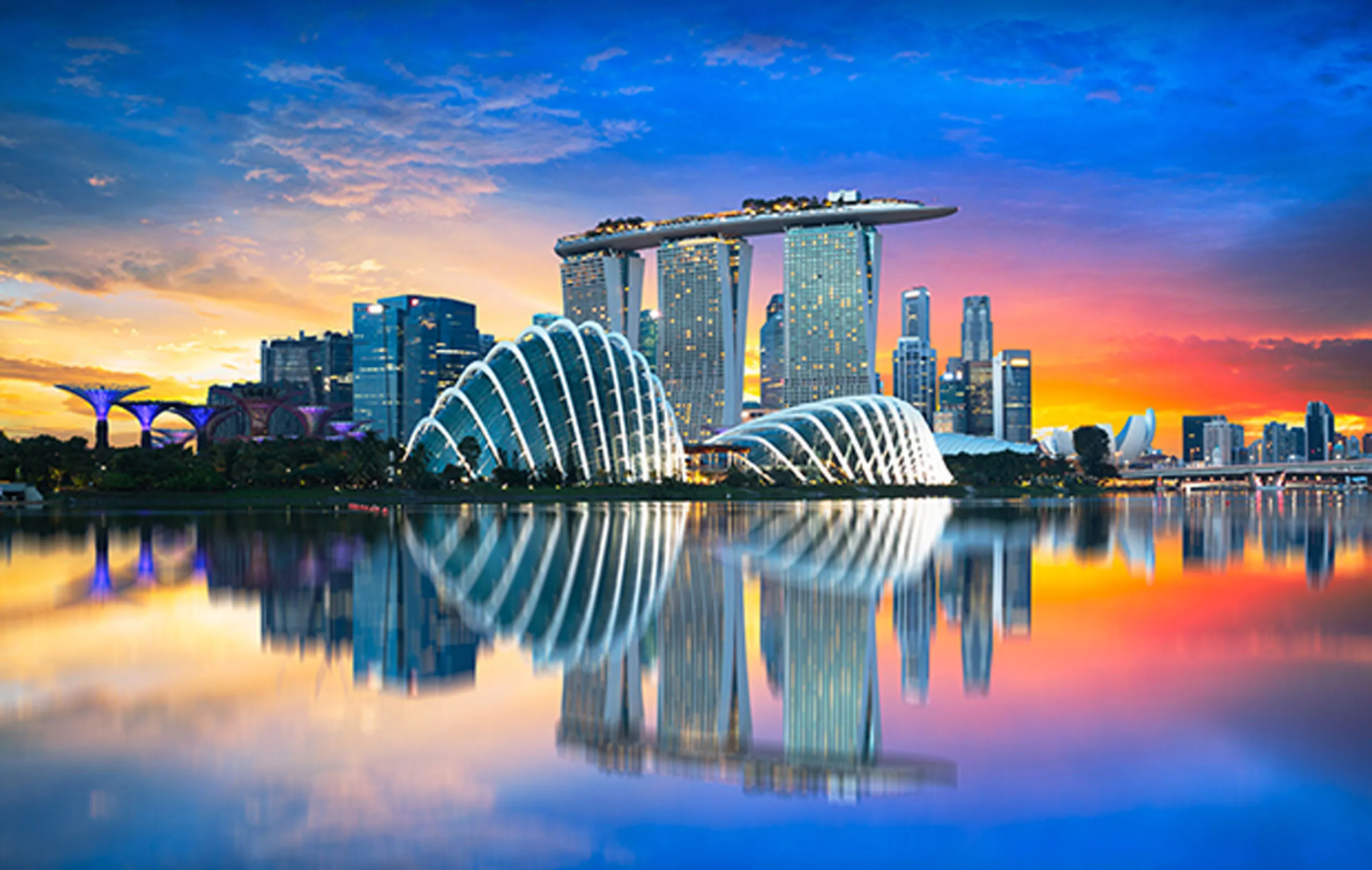 Singapore skyline at sunset featuring Marina Bay Sands and Gardens by the Bay reflected in water.