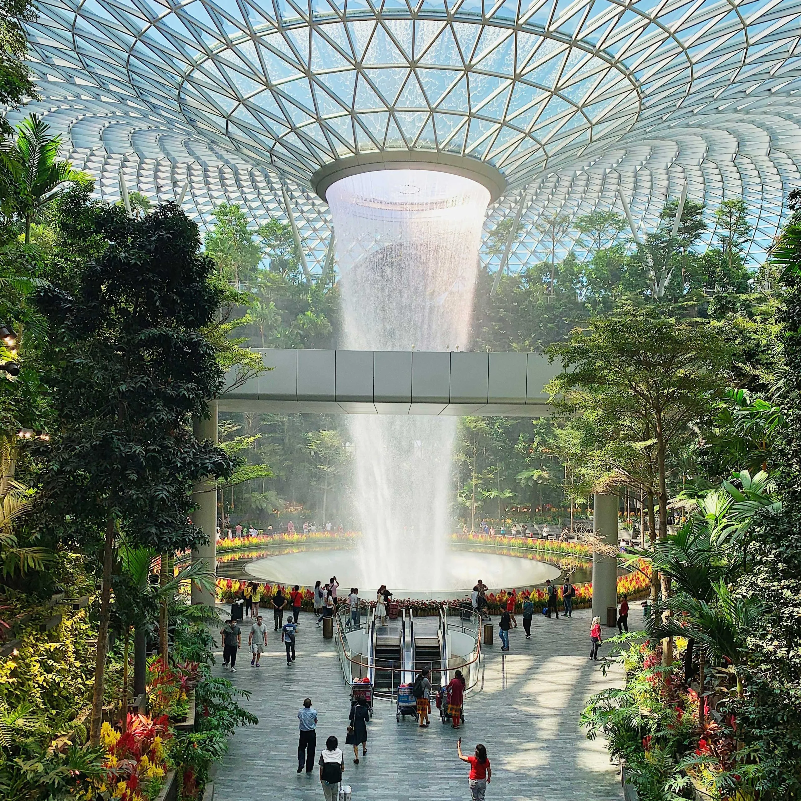 A lush indoor garden with a central waterfall under a glass dome, surrounded by visitors.