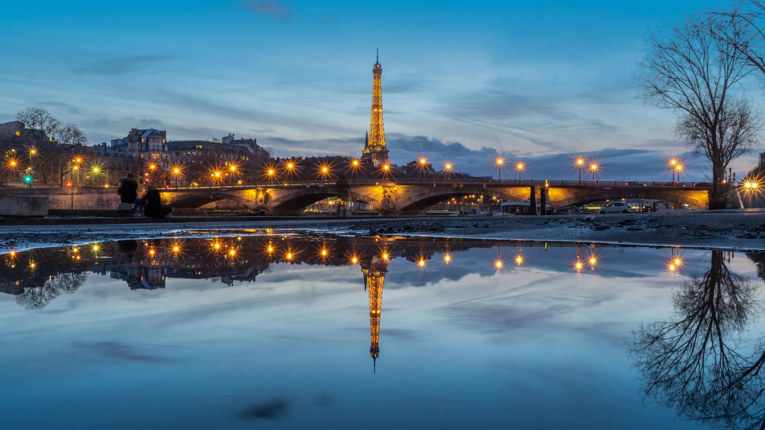 Eiffel Tower and bridge lights reflect in a calm water surface at dusk, framed by trees and cityscape.