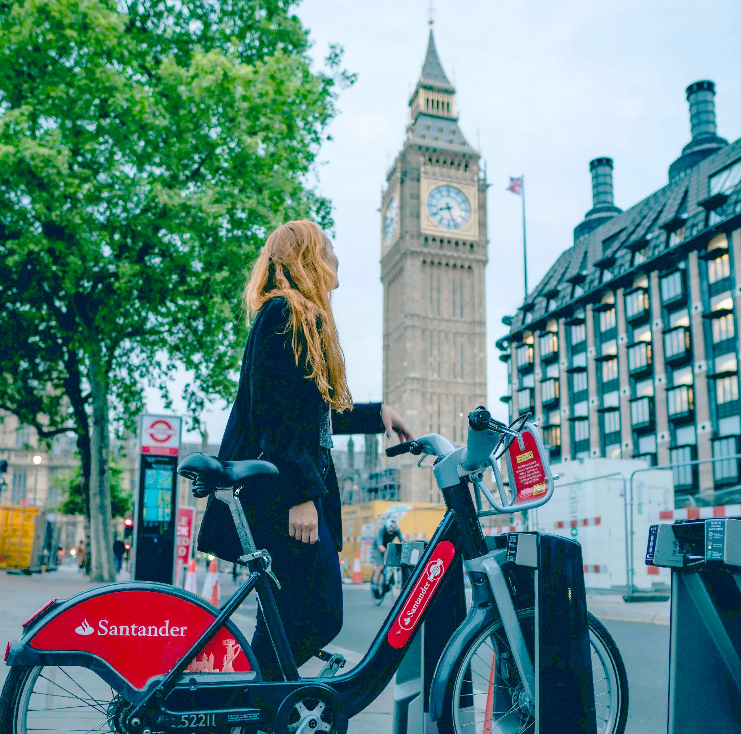 A woman with red hair stands beside a Santander bike near Big Ben in London.