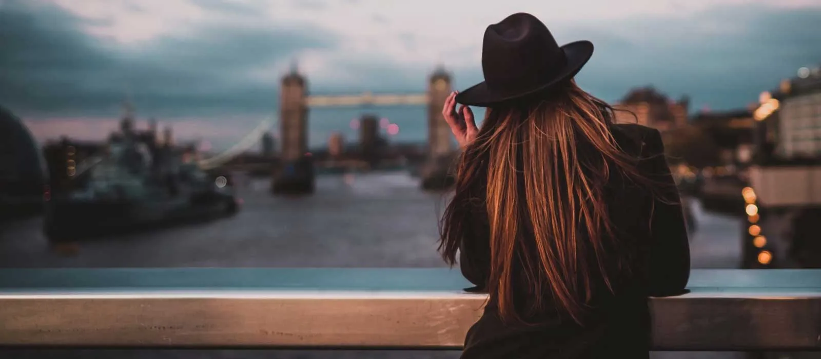 A woman in a black hat overlooks Tower Bridge at dusk, with blurred city lights in the background.