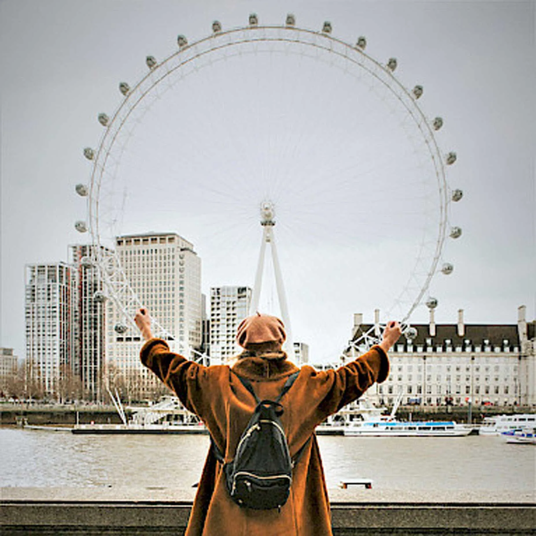 A person in a brown coat faces the London Eye, arms raised, with a cityscape in the background.
